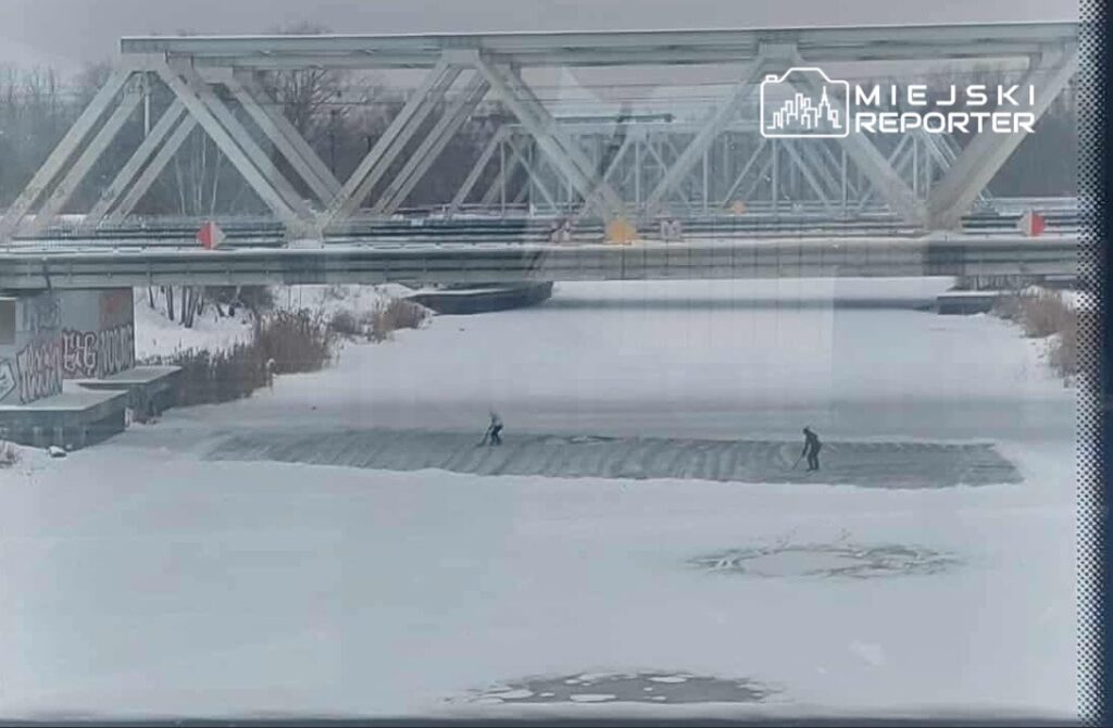Dwóch mężczyzn ślizga się na zamarzniętej rzece pod stalowym mostem, otoczeni śniegiem i lodem.