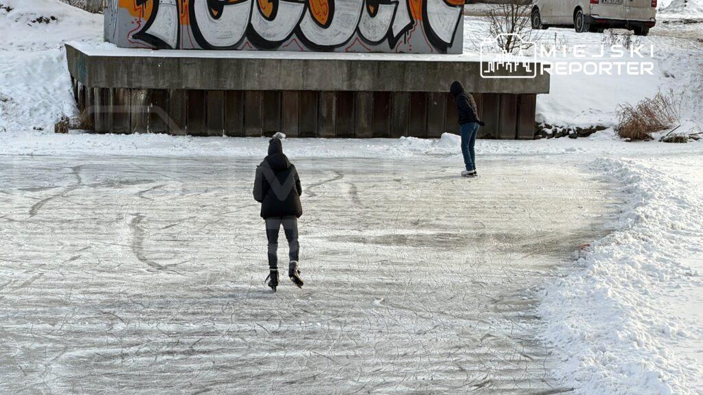 Dwoje łyżwiarzy poruszających się po zamarzniętej tafli lodu w otoczeniu śniegu i graffiti na ścianie.