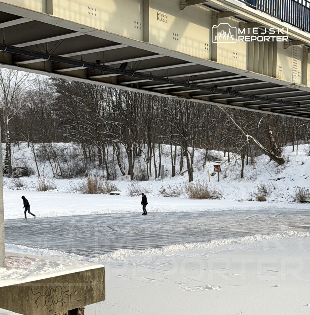 Dwie osoby jeżdżą na łyżwach po zamarzniętej tafli wody pod mostem, otoczonej zaśnieżonymi drzewami.