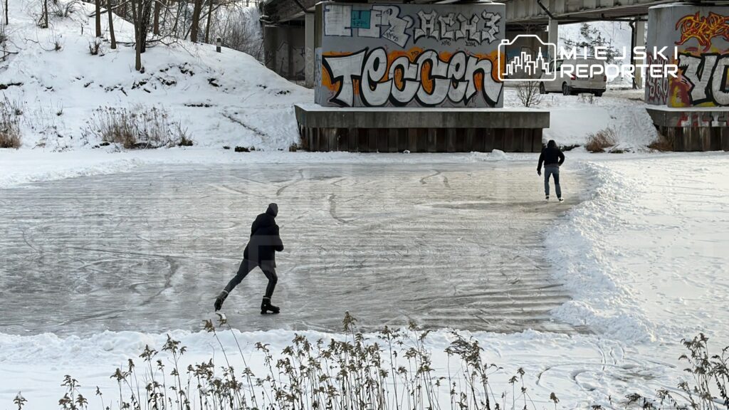 Dwaj łyżwiarze ślizgają się po zamarzniętej tafli lodu w otoczeniu pokrytych śniegiem brzegów i graffiti na murze.