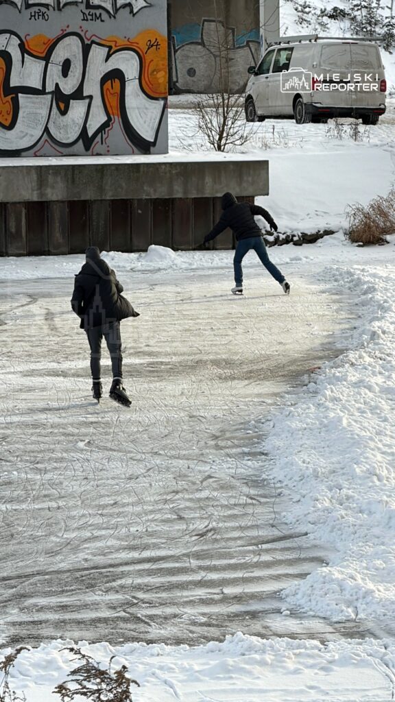 Dwaj chłopcy jeżdżą na łyżwach po zamarzniętym stawie, w tle graffiti na ścianie i zaśnieżony krajobraz.