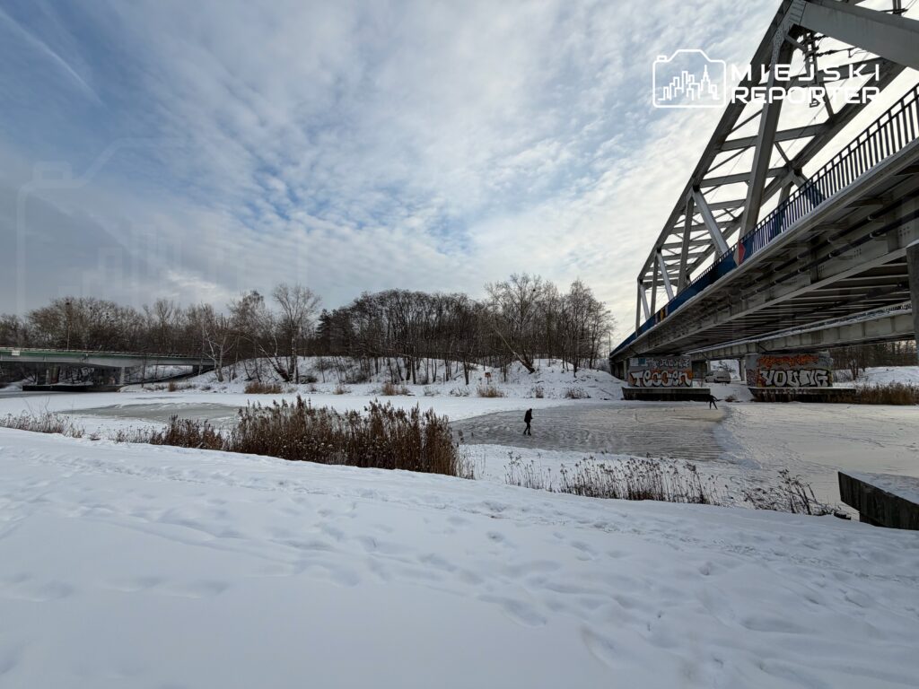 Mężczyzna jeździ na łyżwach na zamarzniętej rzece pod stalowym mostem, otoczony śniegiem i drzewami.