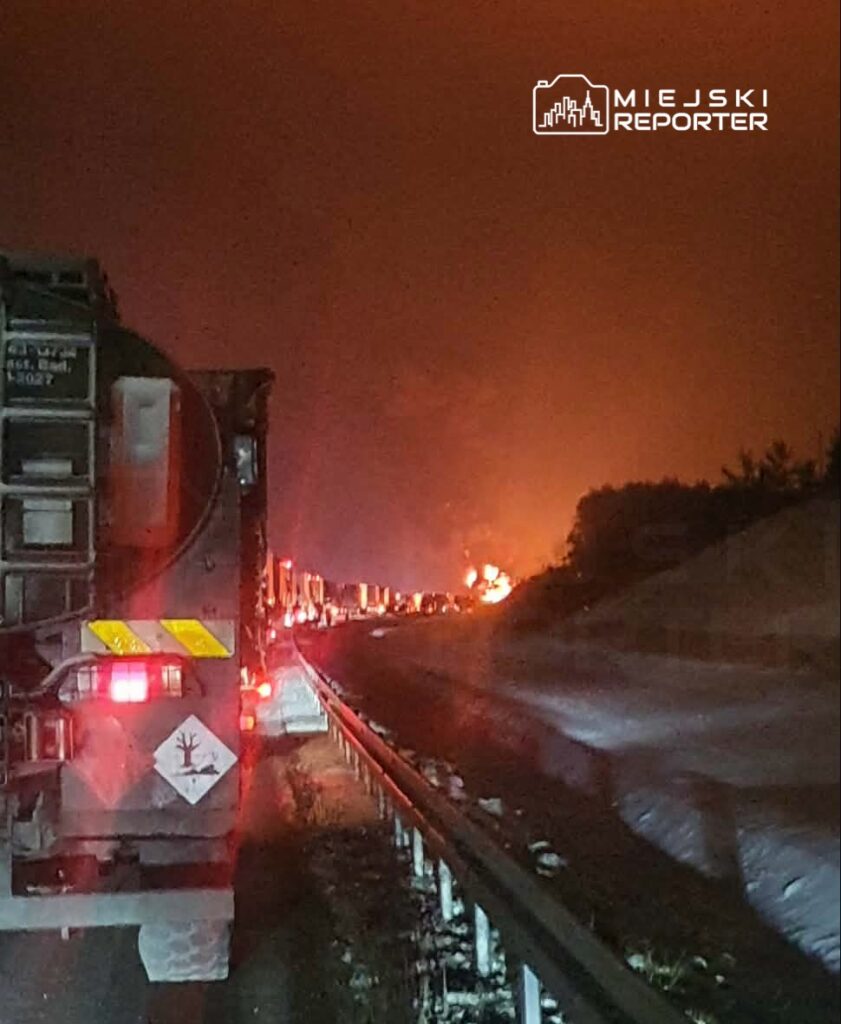 Kierowca ciężarówki stoi w zatorze drogowym, w tle widać ogień i dym na horyzoncie.