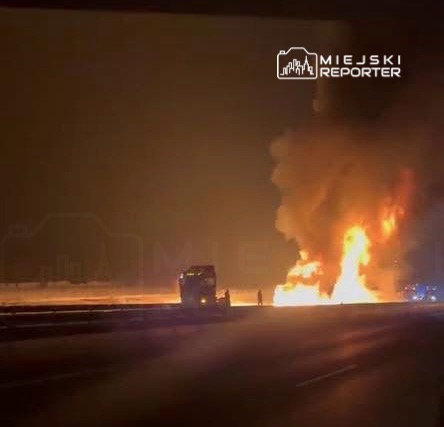 Pojazd ciężarowy płonący na autostradzie, w tle widoczne płomienie i dym.