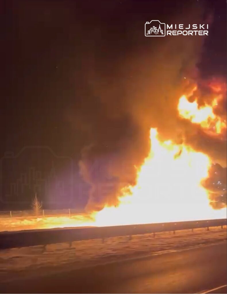 Intensywny pożar z dużymi płomieniami i gęstym dymem widoczny przy drodze w ciemnej scenerii nocnej.