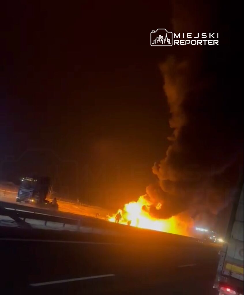 Ogromny pożar pojazdu na autostradzie w nocy, gdzie widać płomienie i gęsty dym, obok stojące ciężarówki.