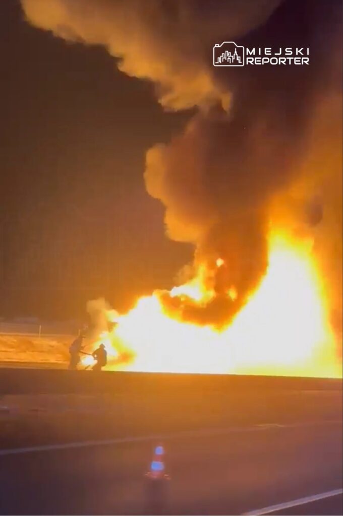 Dwaj strażacy gaszą pożar, który wybuchł na poboczu drogi, otoczony gęstymi, czarnymi kłębami dymu.