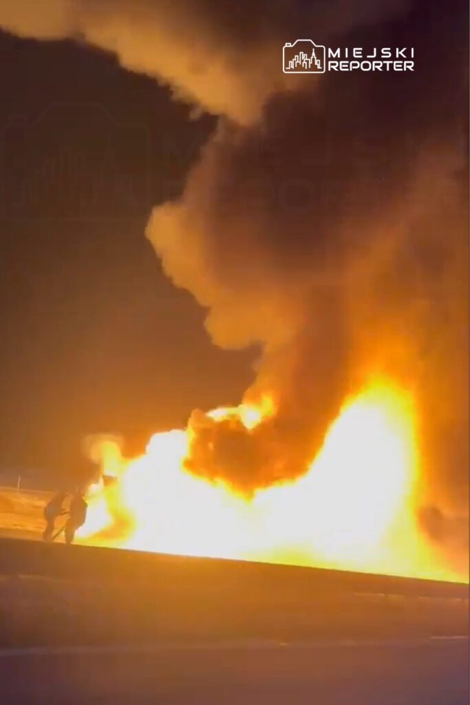 Dwaj mężczyźni w ciemnych ubraniach próbują ugasić płonący pojazd w nocy, otoczeni gęstym dymem.