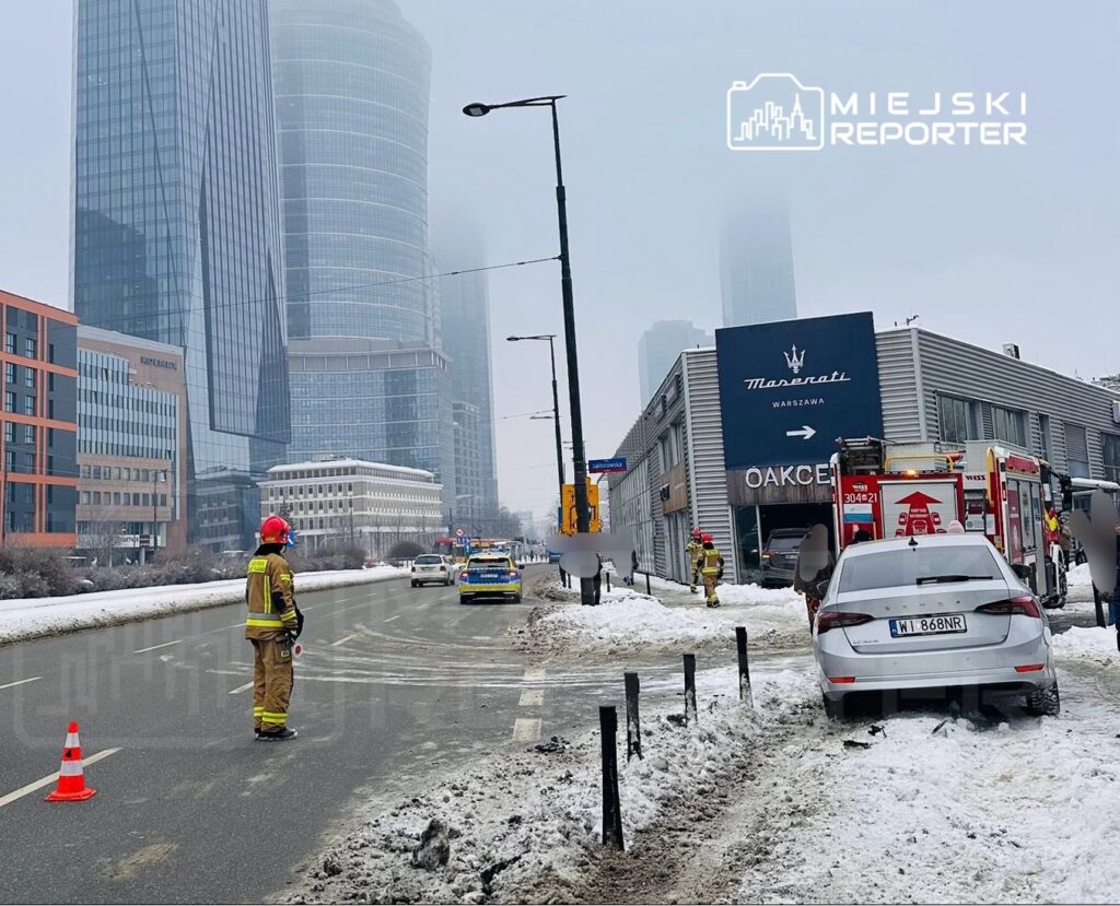 Strażak w hełmie kieruje ruchem na zaśnieżonej ulicy obok uszkodzonego samochodu i wozu strażackiego.