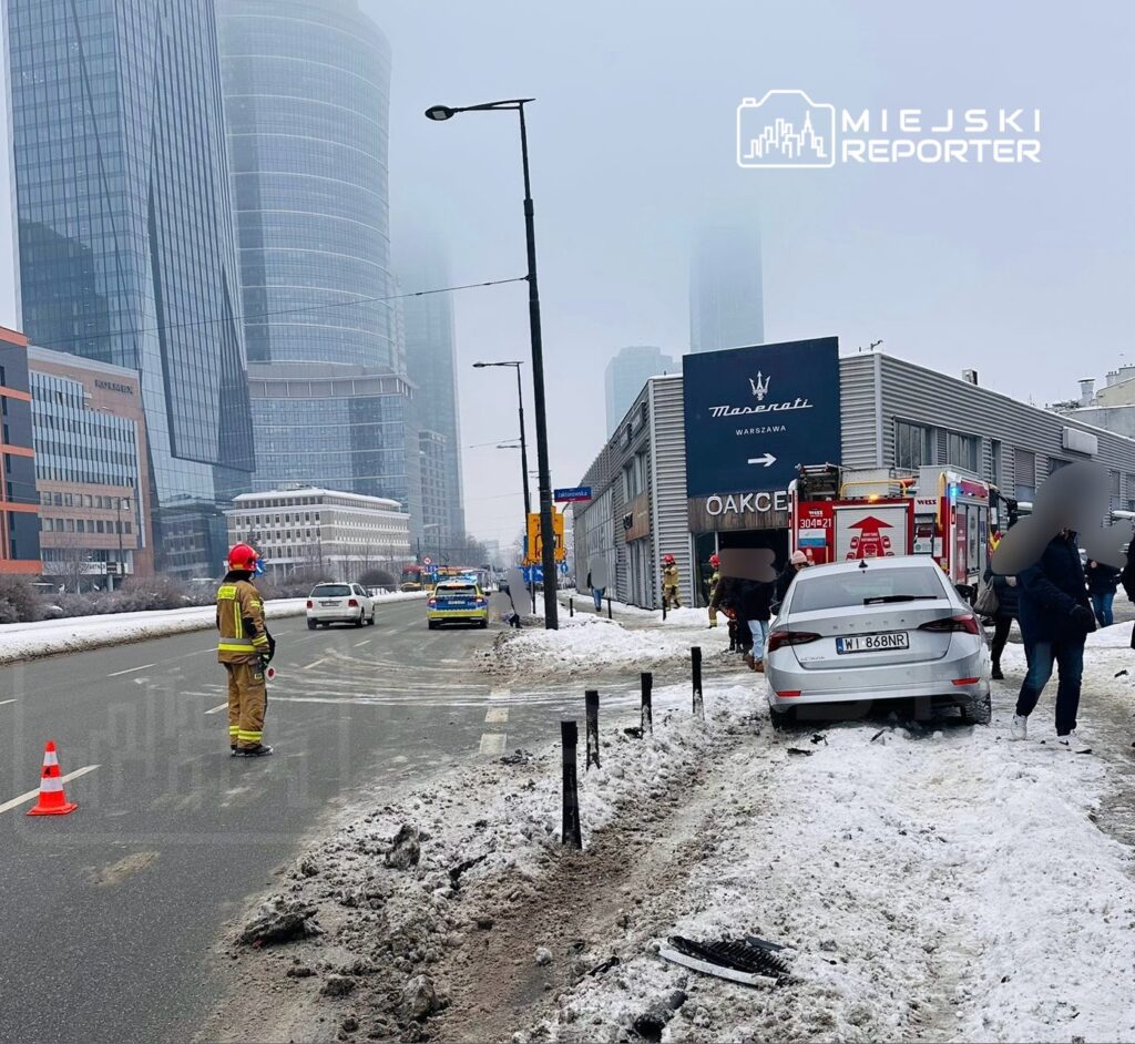 Strażak w hełmie kontrolujący ruch obok uszkodzonego samochodu na zaśnieżonej ulicy w otoczeniu nowoczesnych budynków.