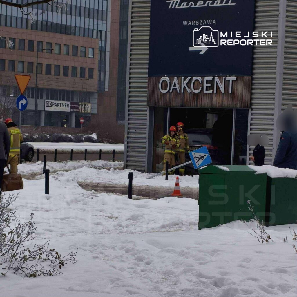 Grupa strażaków w mundurach z hełmami przebywa przed budynkiem z napisem "OAKCENT", obok uszkodzonego znaku drogowym.