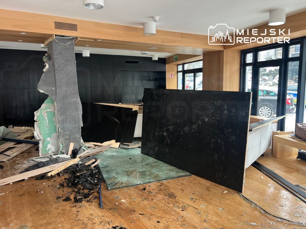 Zniszczona przestrzeń biurowa z usuniętą ścianką działową, rozrzuconymi odpadami budowlanymi i meblami.