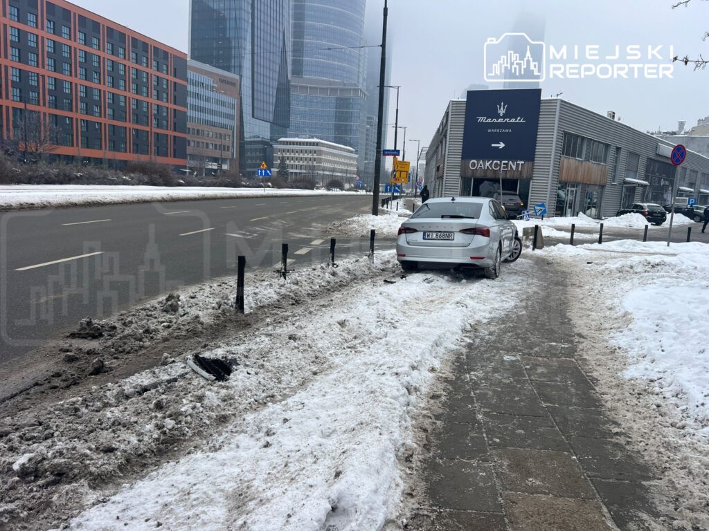 Samochód zaparkowany na chodniku w obszarze miejskim, obok nowoczesnego budynku z reklamą. Wokół leży śnieg.