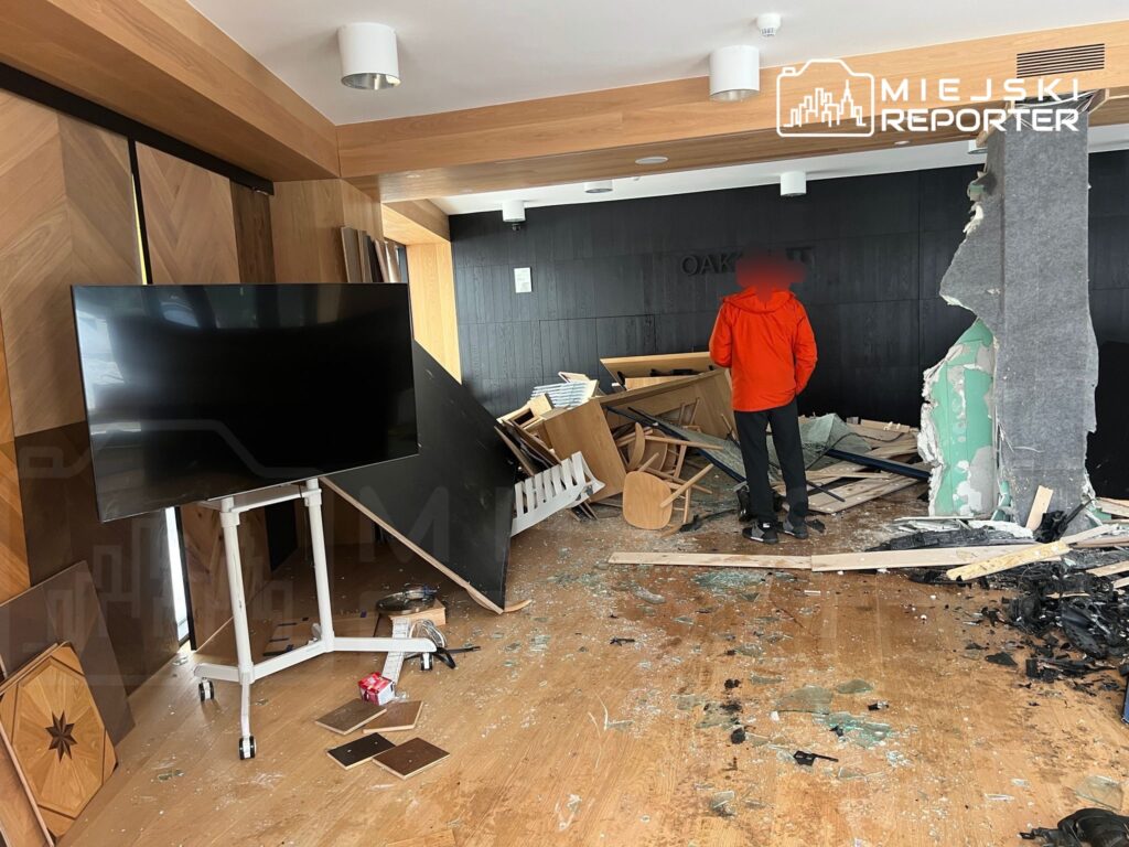 Mężczyzna w czerwonej kurtce stoi w zniszczonym pomieszczeniu z porozrzucanymi meblami i uszkodzonymi ścianami.