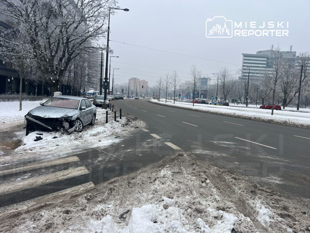 Zniszczony samochód osobowy stoi na chodniku, obok zaśnieżonej jezdni w miejskim otoczeniu.