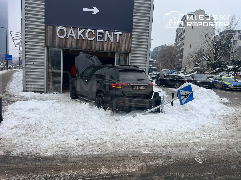 Czarny SUV wjeżdża do budynku z przeszkloną fasadą, obok leży przewrócony znak drogowy na zaśnieżonym chodniku.