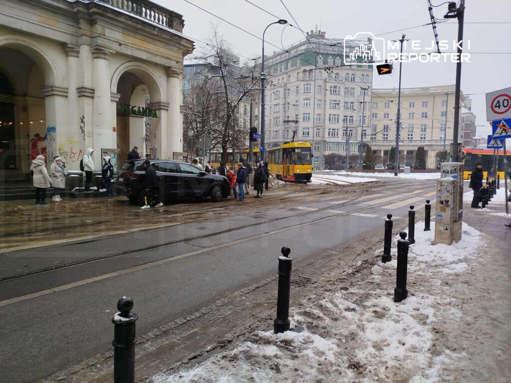 Grupa ludzi w kurtkach czeka na przystanku tramwajowym obok nowoczesnego budynku, w tle widać tramwaj.