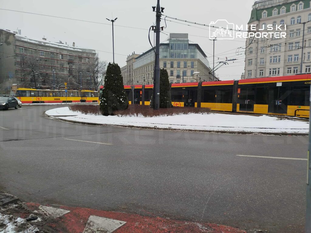 Tramwaj w żółto-czerwonych barwach przejeżdża przez rondo w zimowej scenerii, obok zarośli i budynków.
