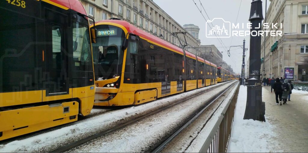 Tramwaje w kolorze żółto-czarnym stoją na ośnieżonym torowisku, obok przechodzą piesi w zimowych ubraniach.