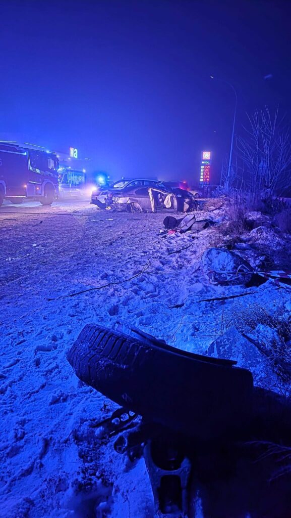 W nocy, w zaśnieżonym otoczeniu, widać zderzenie dwóch samochodów, wokół krążą pojazdy służb ratunkowych z włączonymi sygnałami.
