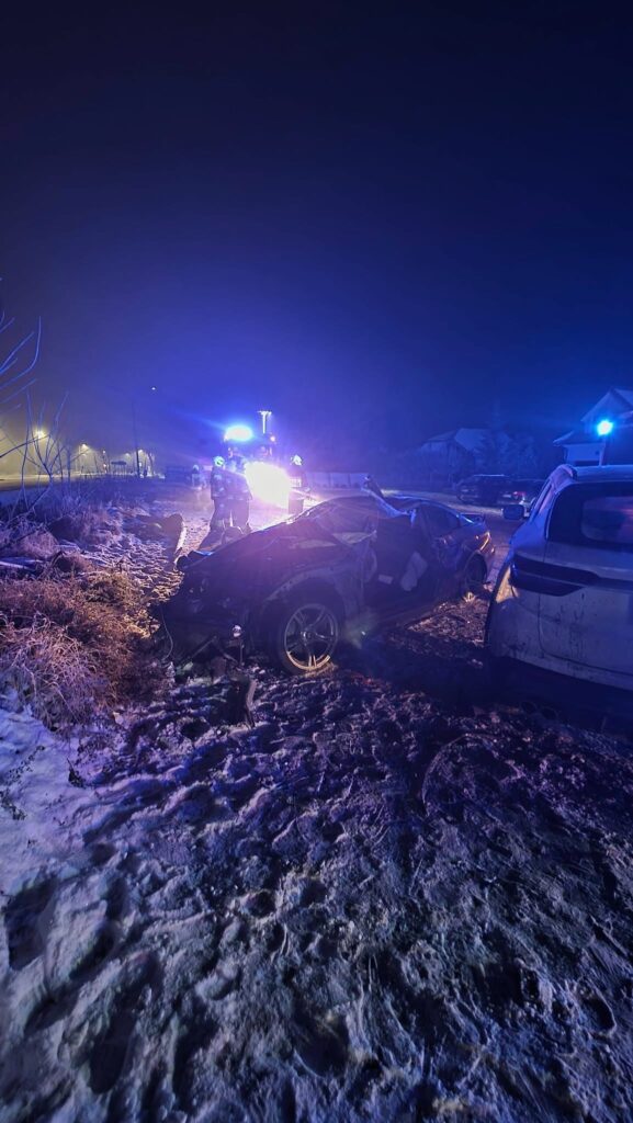Na poboczu drogi znajduje się rozbity samochód, a obok niego działają ratownicy w niebieskich mundurach.
