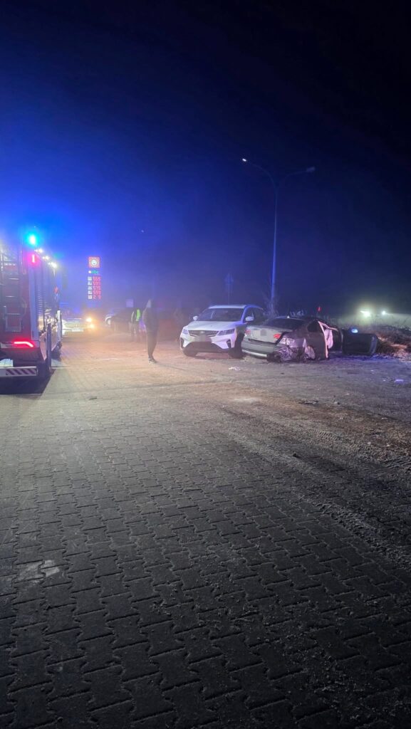 Na drodze w nocy stoją dwa uszkodzone samochody, a obok nich widać straż pożarną oraz osoby w odblaskowych kamizelkach.