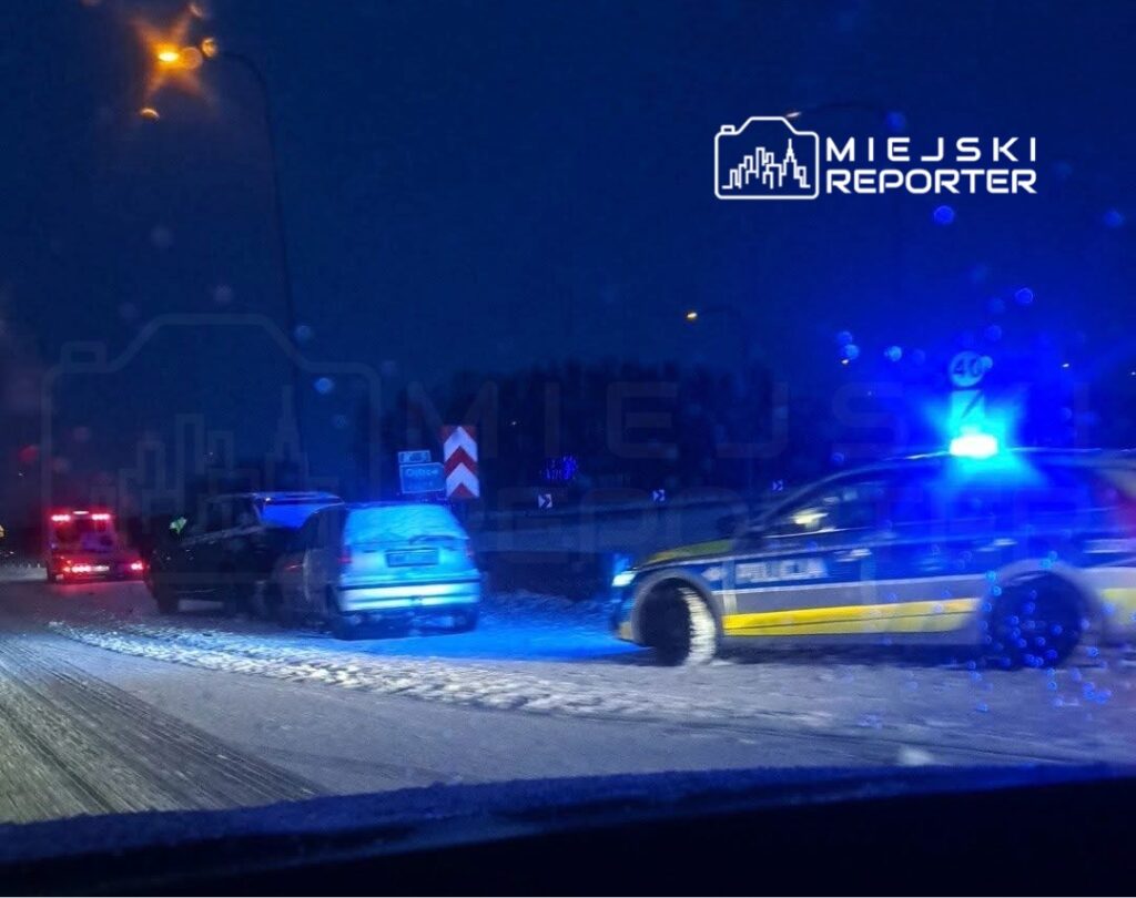 Policyjny radiowóz z włączonymi sygnałami świetlnymi zatrzymany na zaśnieżonej drodze, obok stoi uszkodzony samochód.