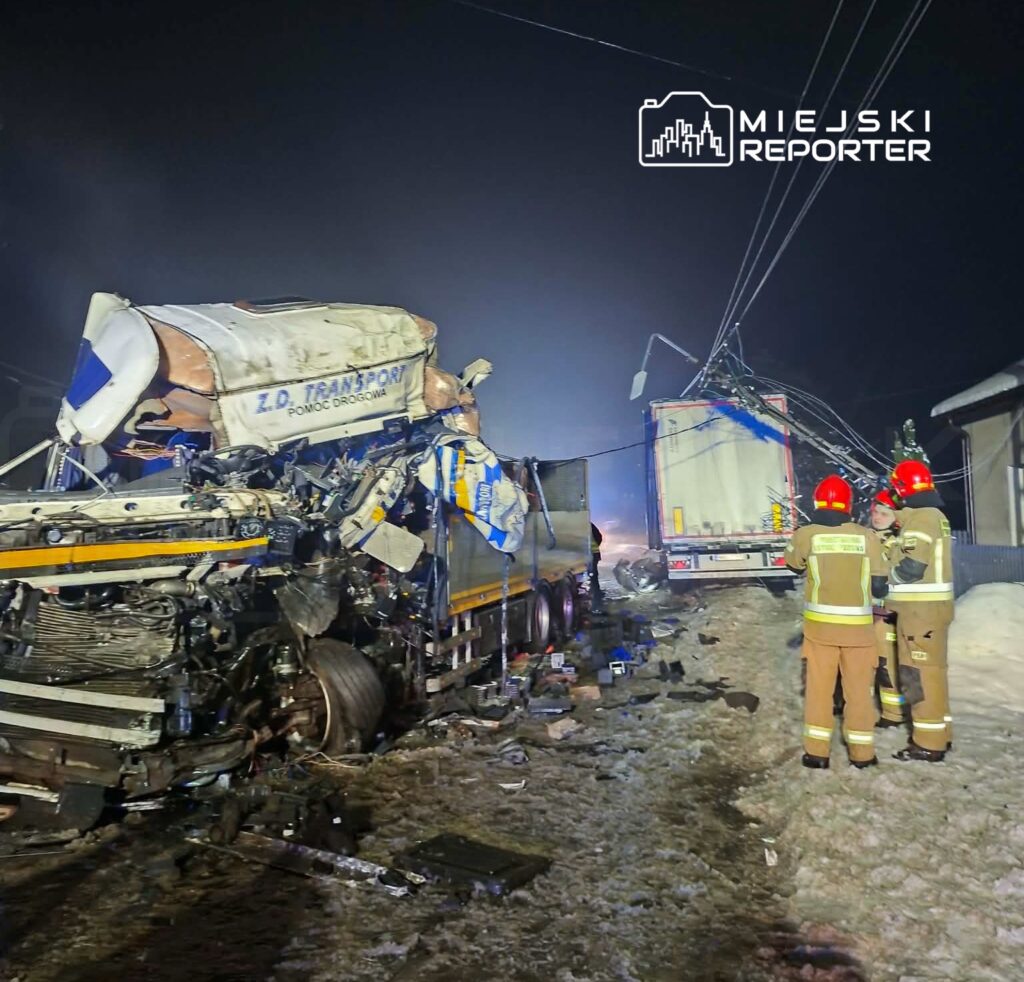 Zniszczone ciężarówki po wypadku drogowym, obok których stoją strażacy w hełmach. W tle widoczna nocna sceneria.