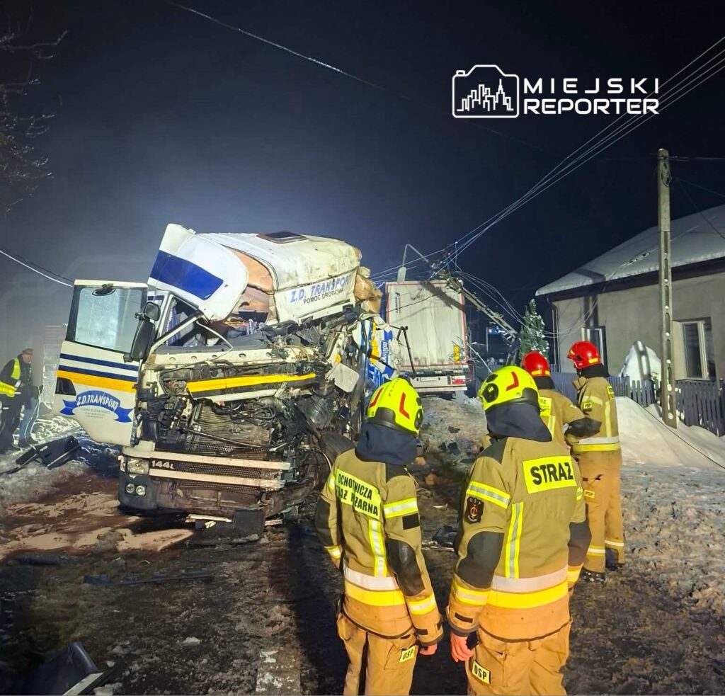 W nocy strażacy w żółtych hełmach oceniają skutki wypadku drogowego z udziałem ciężarówki, w tle widać uszkodzony pojazd.