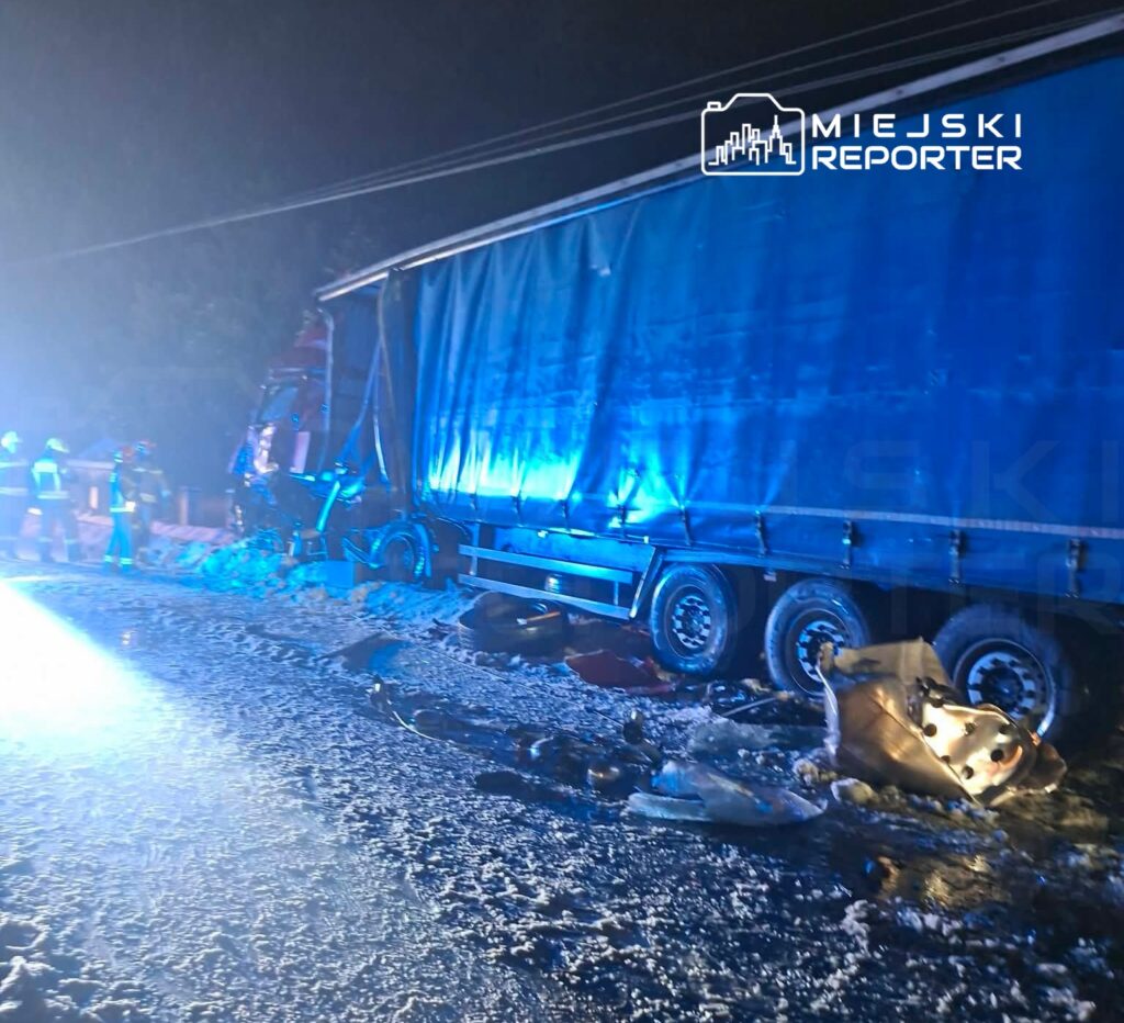 Ciężarówka z uszkodzoną naczepą stoi na zaśnieżonej drodze, a obok pracują strażacy w hełmach.