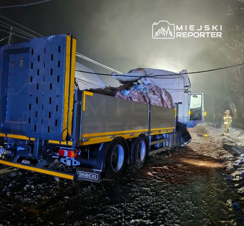 Ciężarówka z załadunkiem materiałów odpadowych stojąca na ośnieżonej drodze, w tle widoczni strażacy.