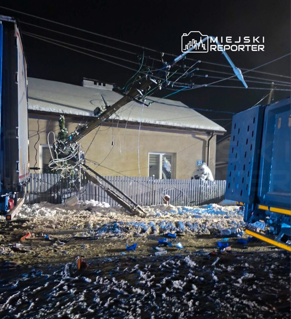 W nocy ciężarówka z uszkodzoną naczepą leży na zaśnieżonej drodze, z przewróconym słupem energetycznym w tle.