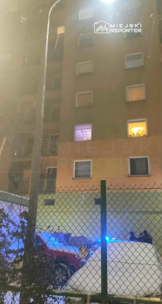 W nocy, w oknie mieszkania na wyższym piętrze, świecą się kolorowe lampki, a poniżej widać zaparkowany samochód pokryty śniegiem oraz radiowóz z włączonymi niebieskimi światłami.