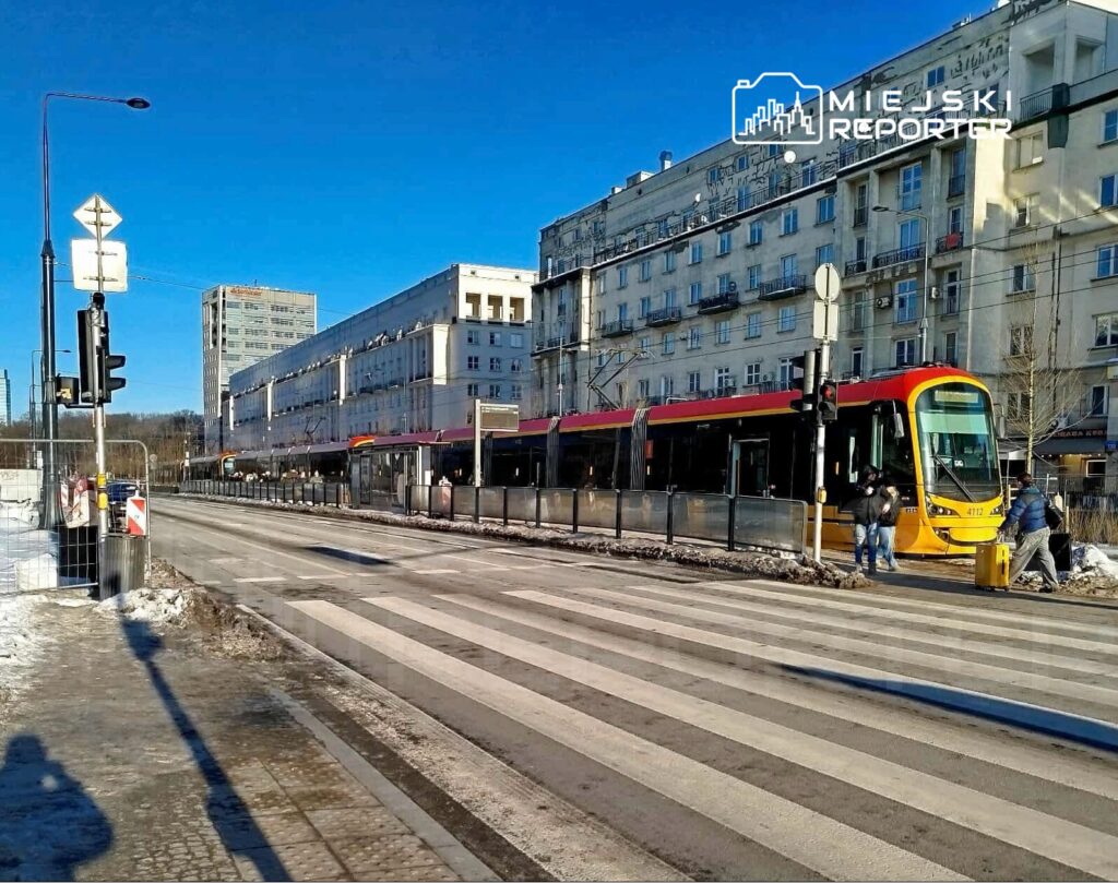 Tramwaj w kolorze żółto-czerwonym zatrzymany na przystanku, obok przejścia dla pieszych w miejskim otoczeniu.