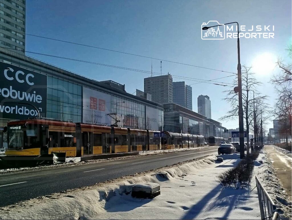 Tramwaj w kolorze żółto-czarnym zatrzymuje się na przystanku przy nowoczesnym budynku z dużymi witrynami.