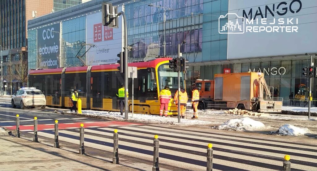 Tramwaj zatrzymany na przystanku, pracownicy w kamizelkach odblaskowych prowadzą prace w pobliżu nowoczesnego biurowca.