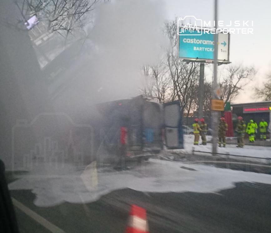Pojazd przewrócony na bok, otoczony przez strażaków w odblaskowych strojach, w tle widoczny znak drogowy.