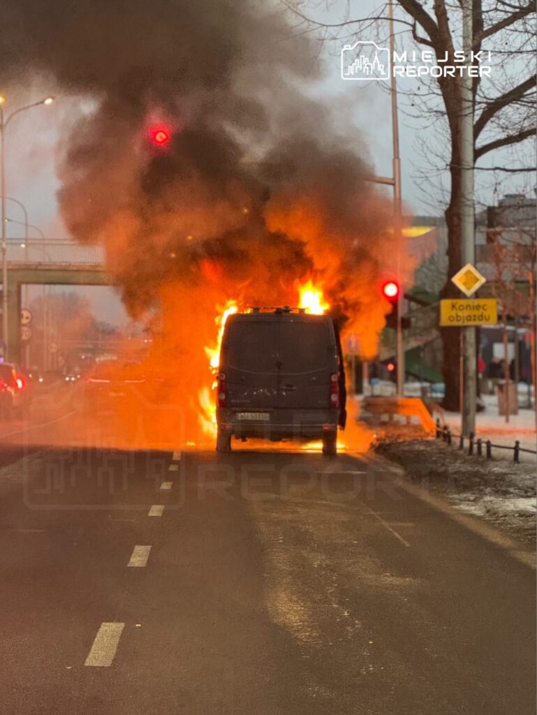 Samochód dostawczy płonie na ulicy, wydobywając gęsty czarny dym, w tle widoczne czerwone światło sygnalizacji.