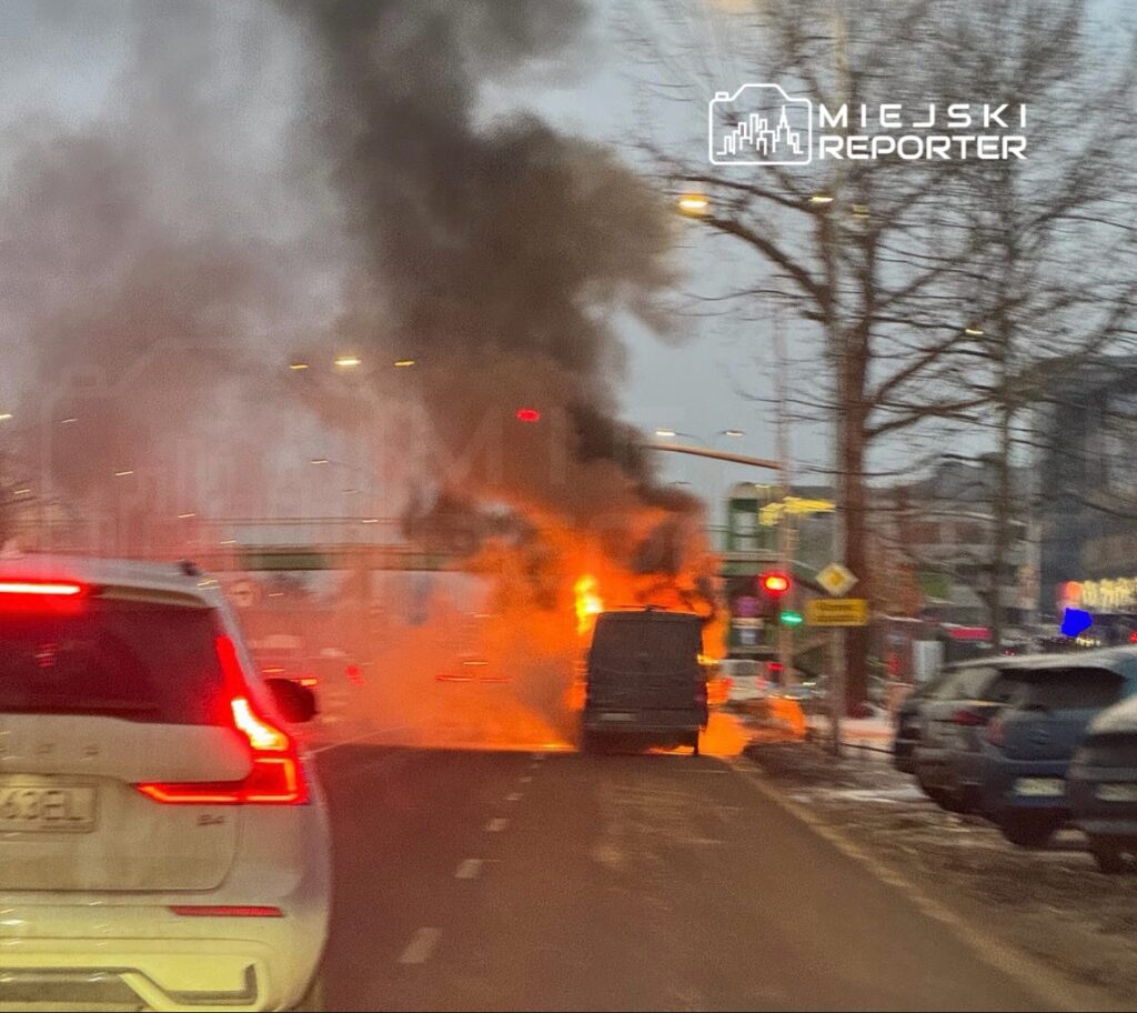 Pojazd płonący na drodze, wydobywający gęsty, czarny dym, w tle widoczne światła uliczne i zaparkowane samochody.