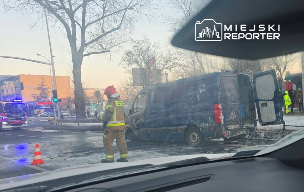 Strażak w hełmie kontroluje gaszenie płonącego furgonu na ulicy, obok widoczny wóz strażacki i grupka ludzi.