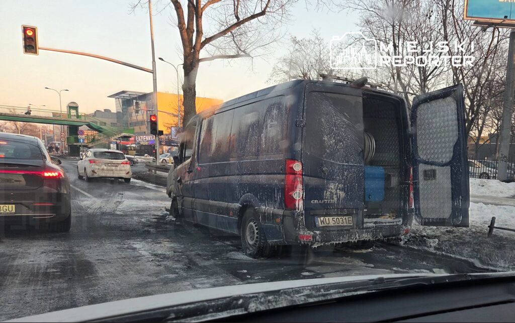 Zniszczony niebieski furgon stoi na poboczu drogi, obok samochodów w ruchu, w tle widoczny jest sygnalizator świetlny.