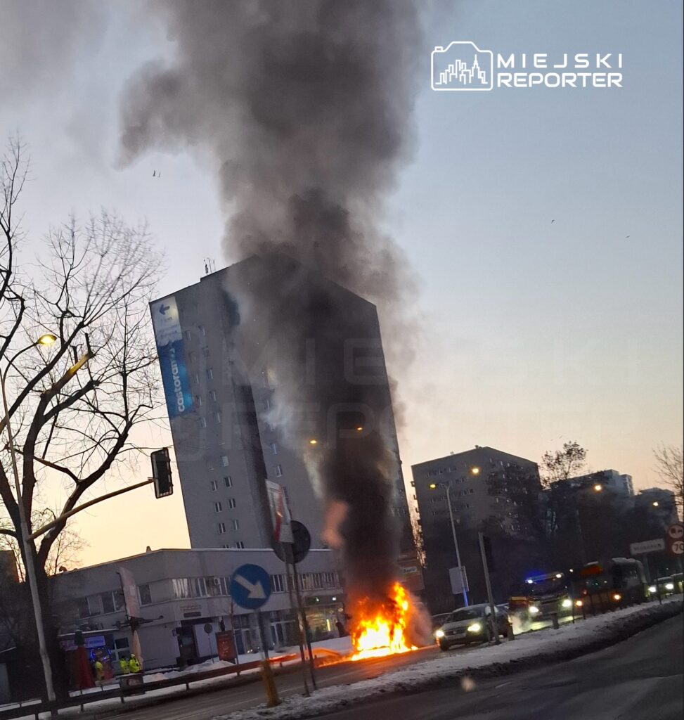 Ogromny słup czarnego dymu unosi się z płonącego pojazdu przy nowoczesnym biurowcu w godzinach porannych.