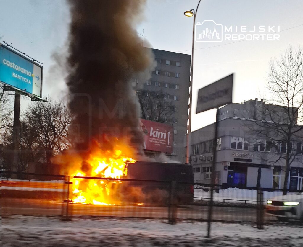 Pojazd płonący na ulicy, wydobywający gęsty dym, w tle budynki i billboard reklamowy.