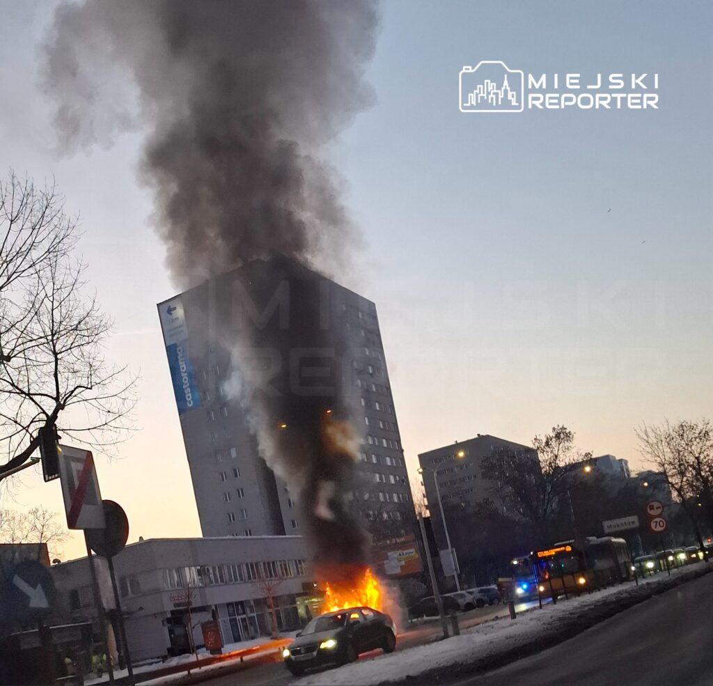 Wysoka wieżowca w tle wydobywa się gęsty, czarny dym, a przed nim zaparkowany samochód płonie.