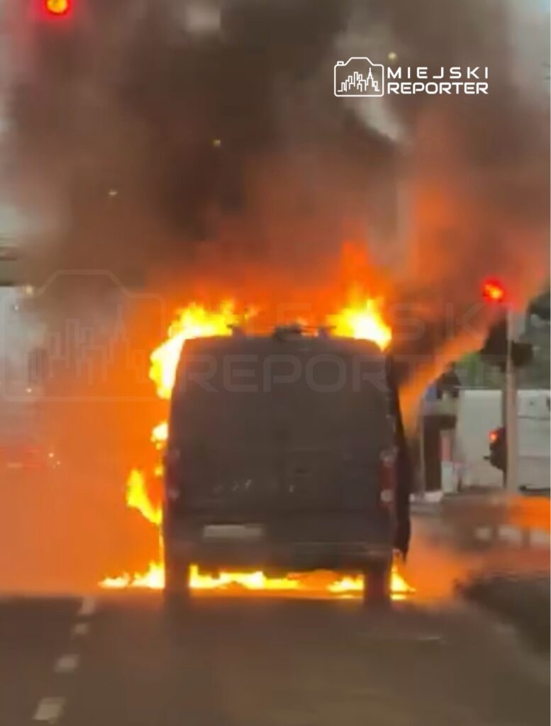 Furgonetka płonąca na drodze, z ogniem wydobywającym się z tyłu i dymem unoszącym się w powietrzu.