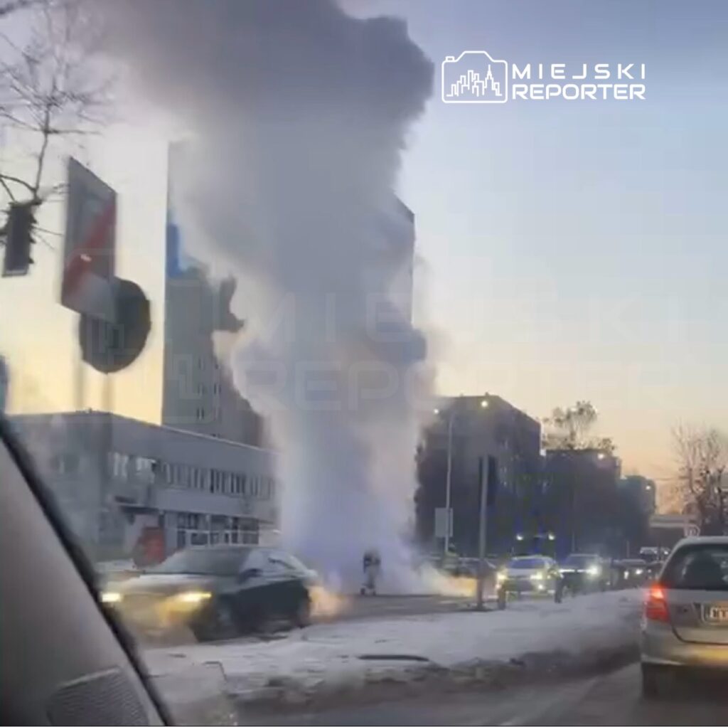 Wysoka chmura pary wydobywająca się z rury na skrzyżowaniu, w tle nowoczesne budynki i ruch uliczny.