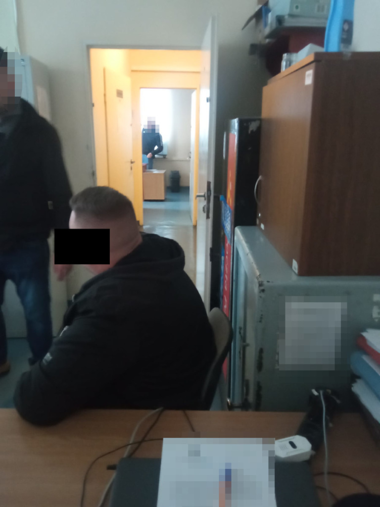 Mężczyzna w czarnej kurtce siedzi przy biurku, podczas gdy inna osoba stoi w drzwiach pokoju o żółtych ścianach.