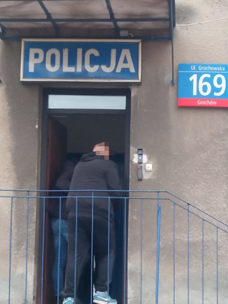 Mężczyźni w ciemnych ubraniach wchodzą do budynku z oznaczeniem "POLICJA" na ścianie.