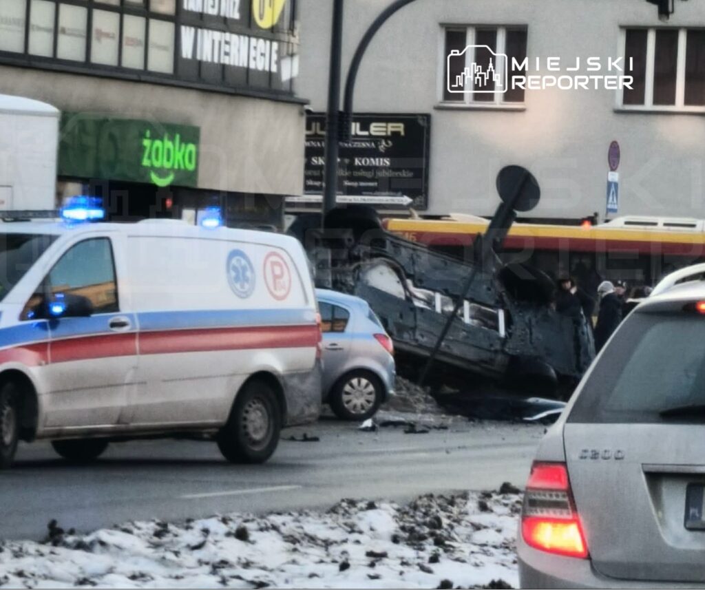 Wielki pojazd przewrócony na bok na skrzyżowaniu, obok zatrzymana karetka pogotowia i zablokowane samochody.