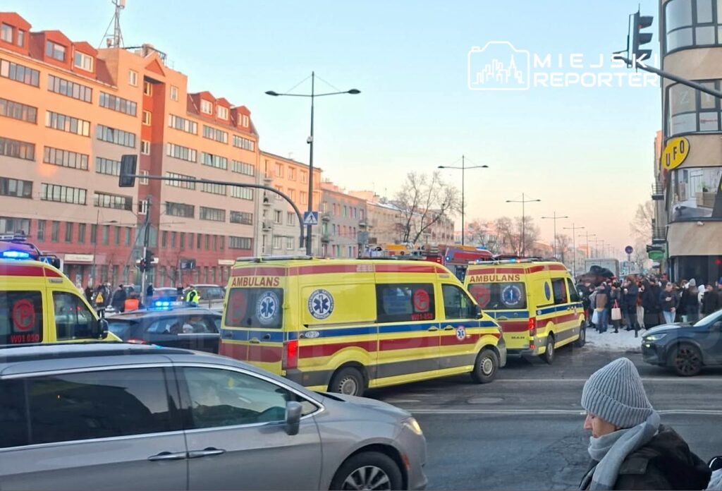 Karetkę pogotowia z włączonymi sygnałami świetlnymi parkują w pobliżu grupy ludzi na chodniku w miejskiej scenerii.